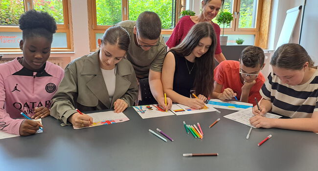 7 Kinder sitzen/stehen an einem Tisch und gestalten ein Arbeitsblatt farbig