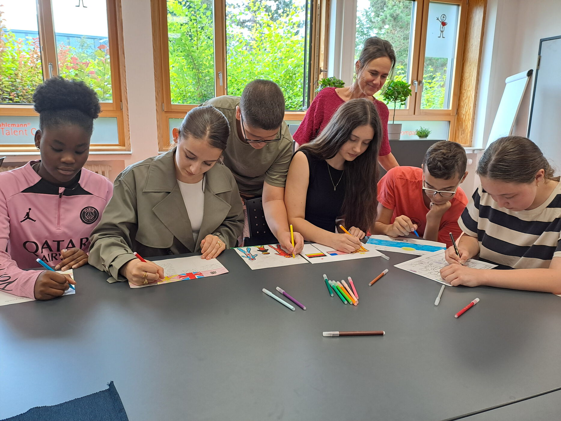 7 Kinder sitzen/stehen an einem Tisch und gestalten ein Arbeitsblatt farbig